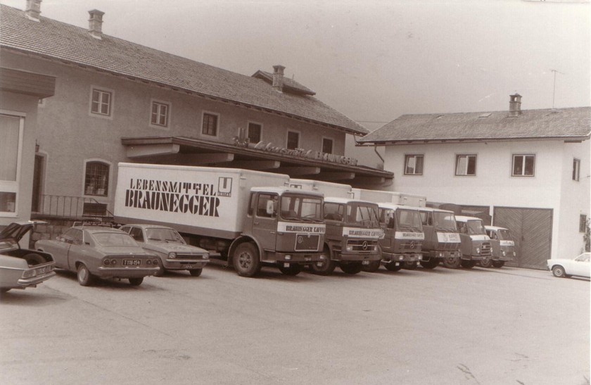 Schwarzweißfoto: Braunegger-Lastwagen und Pkw vor dem Firmengebäude